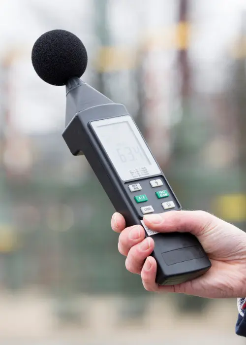 Mano sosteniendo un sonómetro portátil con parabrisas de espuma mientras mide el nivel de ruido en exterior.