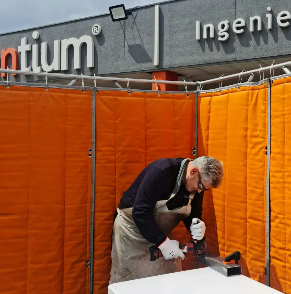Operario utilizando herramienta eléctrica en una estación de trabajo, rodeado por paneles acústicos flexibles de color naranja montados en una estructura tubular para contener el ruido.