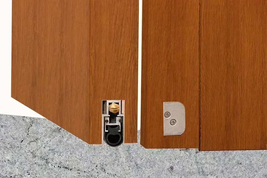 sello acústico automático empotrado (bulbo EPDM y actuador lateral) mostrando cierre hermético sobre piso; parte del sistema de sellos acústicos 360° para control de ruido y filtraciones.