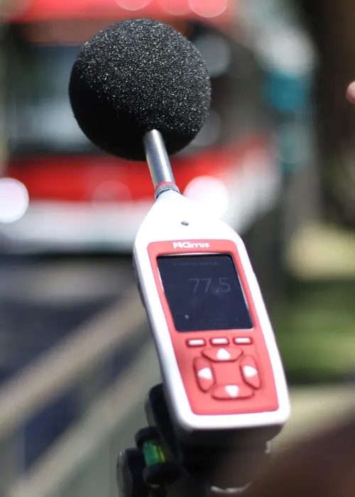 Sonómetro clase 2 montado en trípode midiendo ruido de tránsito (LAeq/Lmax) frente a un bus, registro con datalogger para control acústico urbano.