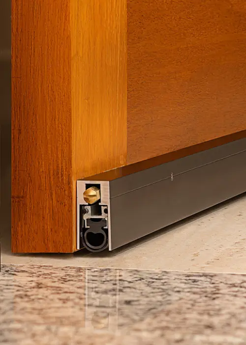 sello acústico automático de caída instalado en el canto inferior de una puerta de madera, parte del sistema de sellos acústicos 360° para control de ruido, polvo y hermeticidad.